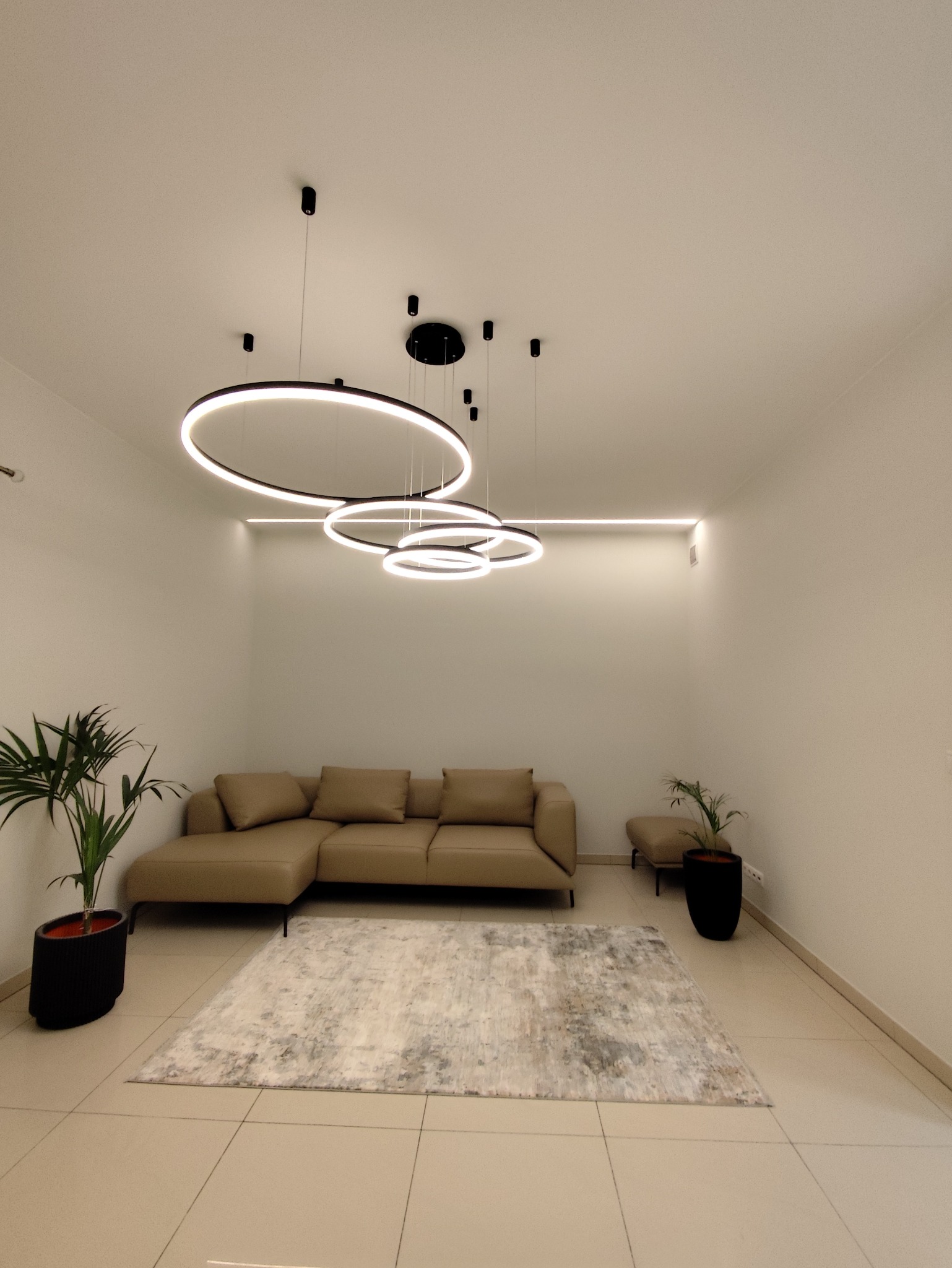 Nowoczesny salon z beżową sofą, wzorzystym dywanem i designerskimi lampami w kształcie okręgów. Minimalistyczne wnętrze z roślinami w czarnych donicach.