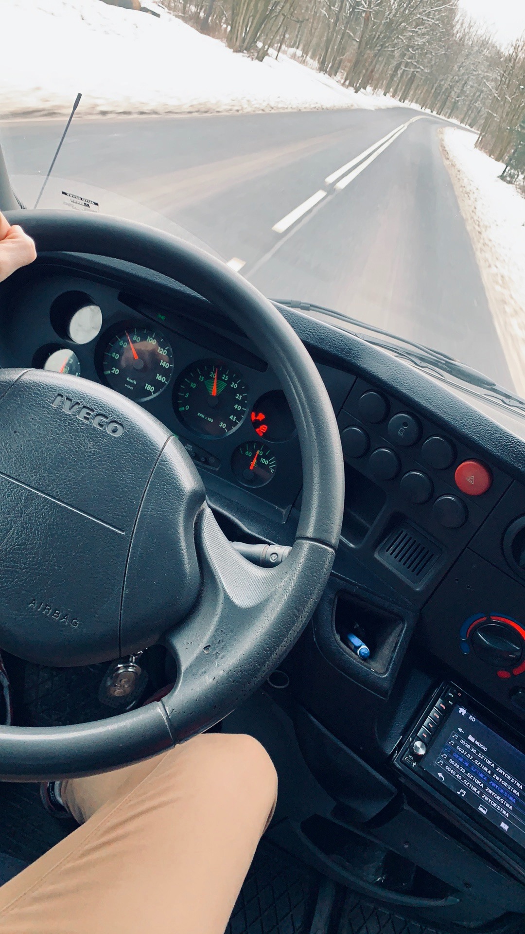 Wnętrze kabiny samochodu ciężarowego Iveco podczas jazdy po ośnieżonej drodze, widok z perspektywy kierowcy na deskę rozdzielczą, kierownicę z napisem AIRBAG i włączone radio. Widoczny fragment...