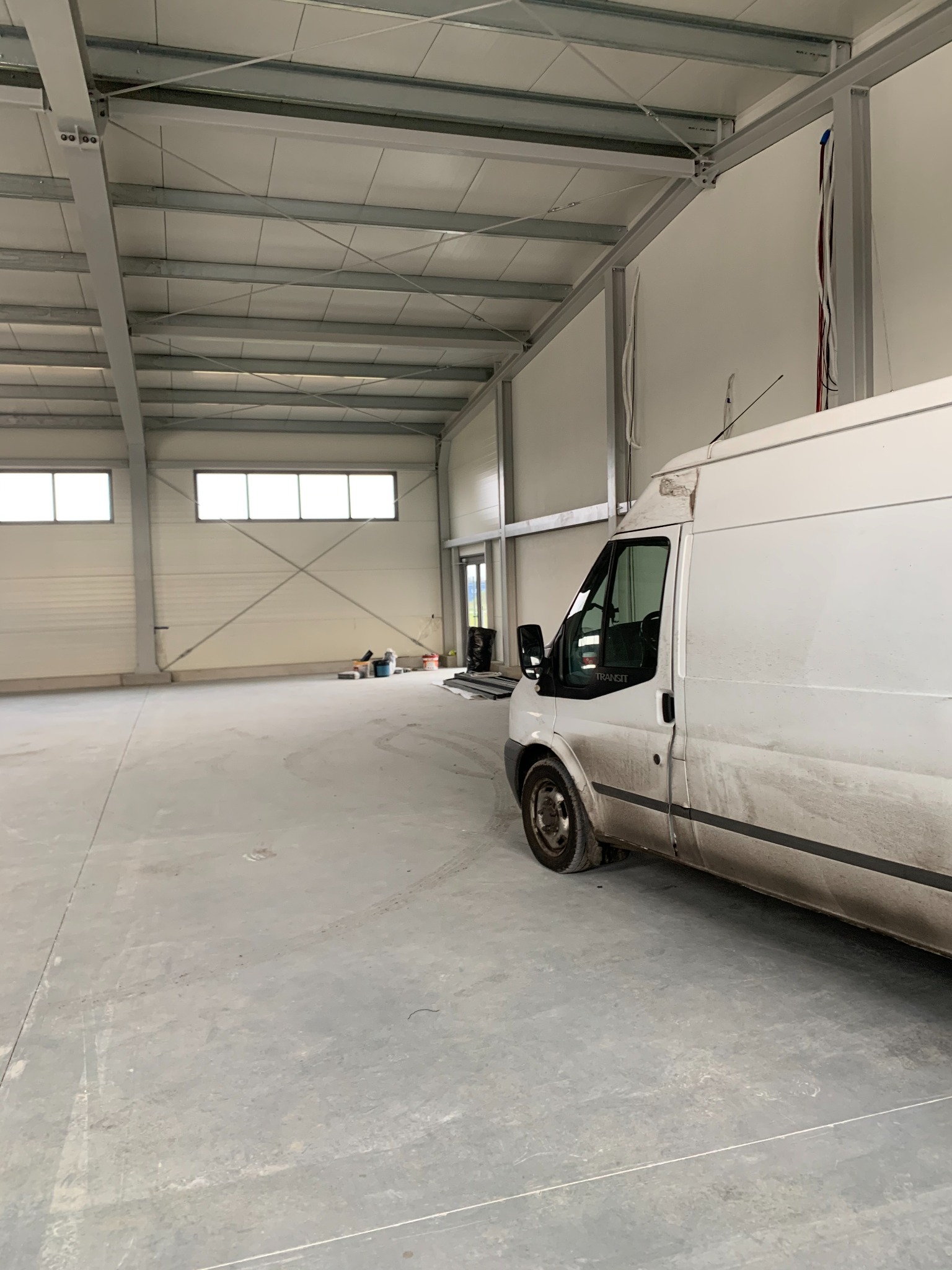 Biały, lekko zabrudzony bus marki Ford Transit zaparkowany wewnątrz nowej, przestronnej hali magazynowej z widoczną konstrukcją stalową dachu i białymi ścianami.