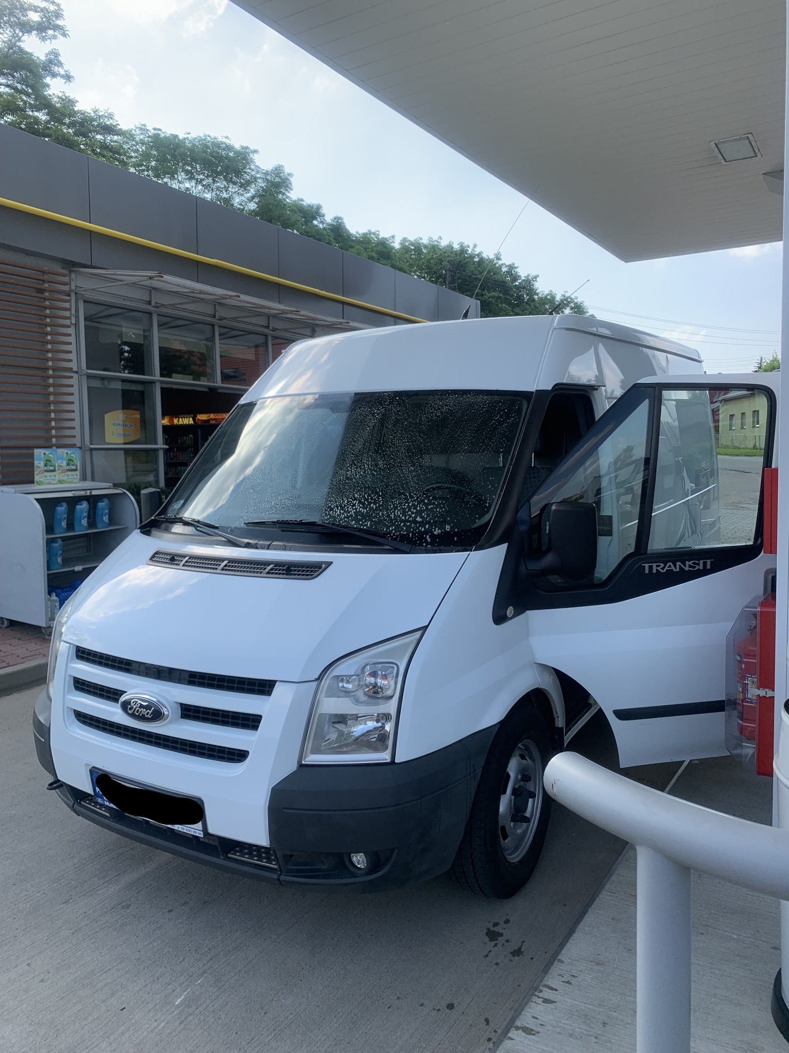 Biały Ford Transit z otwartymi drzwiami, myty na stacji benzynowej w Komornikach, widoczne krople wody na przedniej szybie.