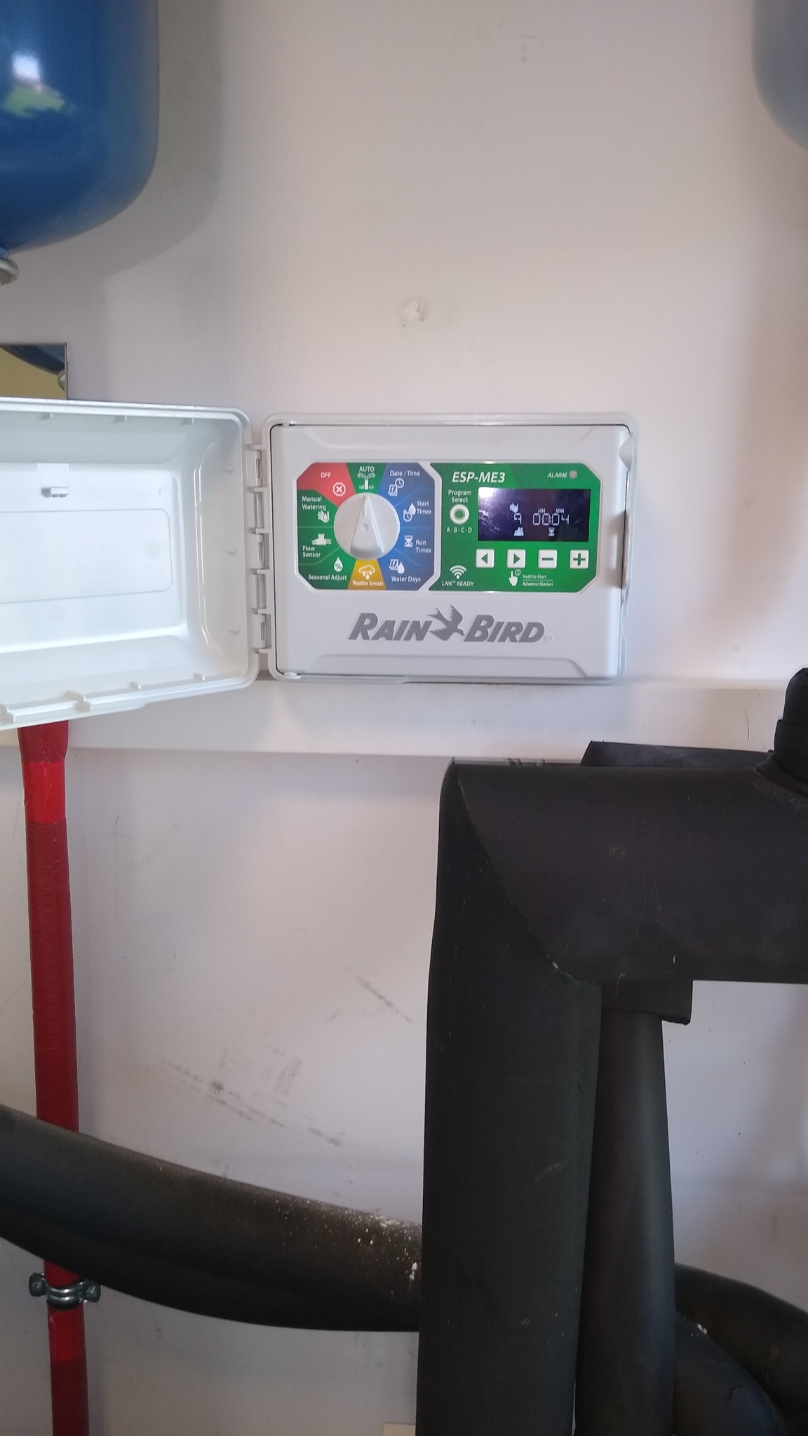 Sterownik nawadniania Rain Bird ESP-ME3 z otwartą obudową, widoczne ustawienia programów i wyświetlacz LCD, zamontowany na ścianie w pomieszczeniu technicznym, z widocznymi instalacjami...