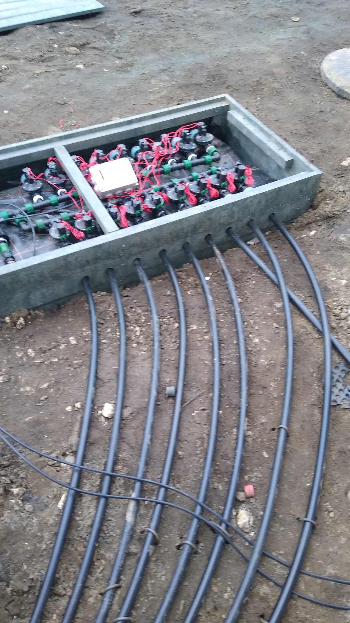 Skrzynka z rozdzielaczem rur systemu nawadniania, widoczne liczne złączki, węże i przewody, całość umieszczona w ziemi.