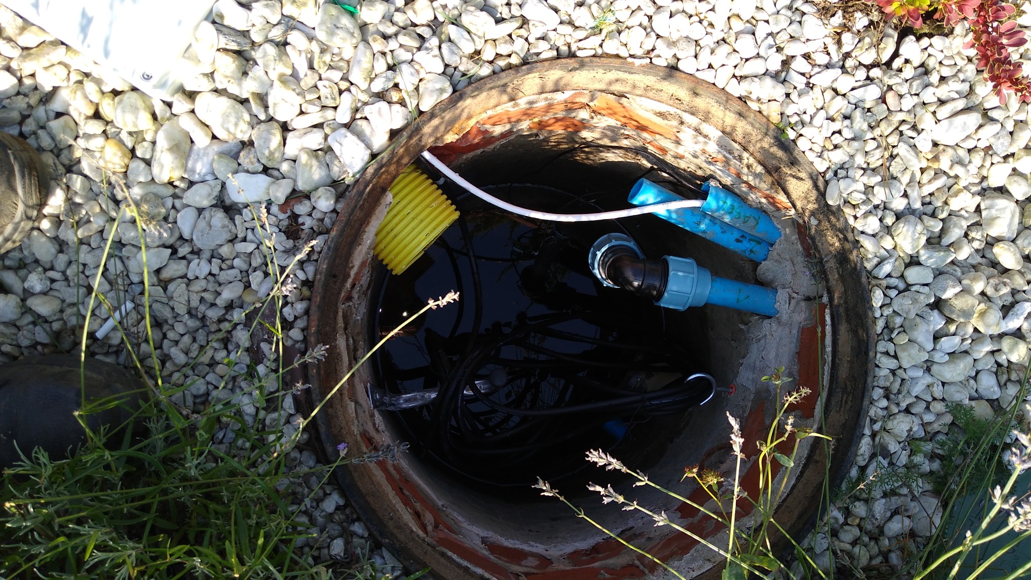 Studzienka rewizyjna w Chałupkach z widocznymi przewodami i elementami instalacji wodnej, otoczona kamieniami i roślinnością.