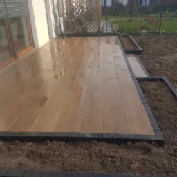 Nowoczesny taras z płytek imitujących drewno, obrzeże z ciemnej kostki, widok na ogród i zabudowę jednorodzinną.