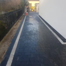 Świeżo ułożona, czarna kostka brukowa z jasnoszarymi obrzeżami, widoczna ubijarka w oddali i pracownik w tle.