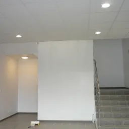 Wnętrze biurowe po odświeżeniu: pomalowane na biało ściany, szare płytki na podłodze, schody z metalową barierką, widoczny wałek do malowania i butelka wody.