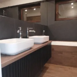 Nowoczesna łazienka z dwoma białymi, prostokątnymi umywalkami nablatowymi, chromowanymi bateriami, ciemnoszarą płytką na ścianach, drewnianymi oknami z żaluzjami oraz drewnopodobną płytką...