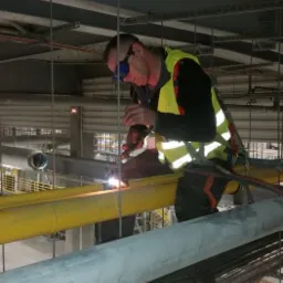 Spawacz w okularach ochronnych i kamizelce odblaskowej, zabezpieczony uprzężą, spawa rurę w instalacji przemysłowej pod sufitem.