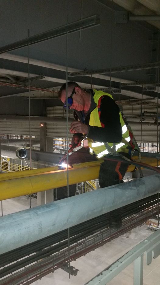 Spawacz w okularach ochronnych i kamizelce odblaskowej, zabezpieczony uprzężą, spawa rurę w instalacji przemysłowej pod sufitem.