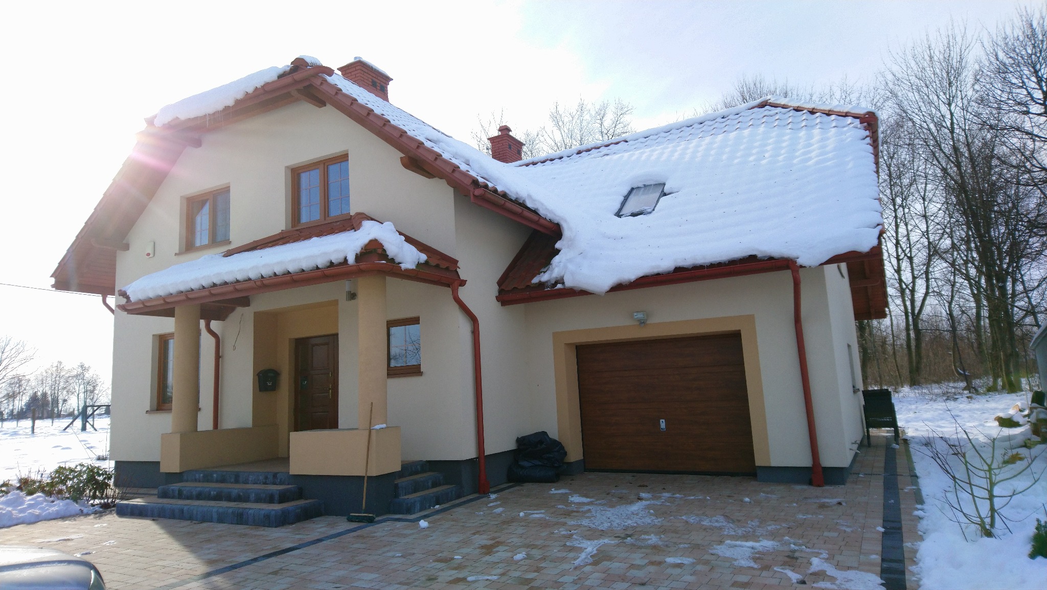 Dom jednorodzinny z beżową elewacją, czerwoną dachówką i ośnieżonym dachem, widoczny od frontu w zimowej scenerii.