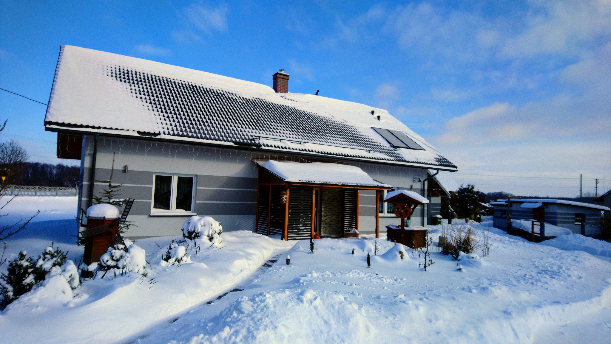 Dom jednorodzinny w zimowej scenerii, pokryty śniegiem, z widoczną elewacją w odcieniach szarości z poziomymi pasami, ciemna dachówka, małe panele solarne na dachu, drewniana weranda i studnia...
