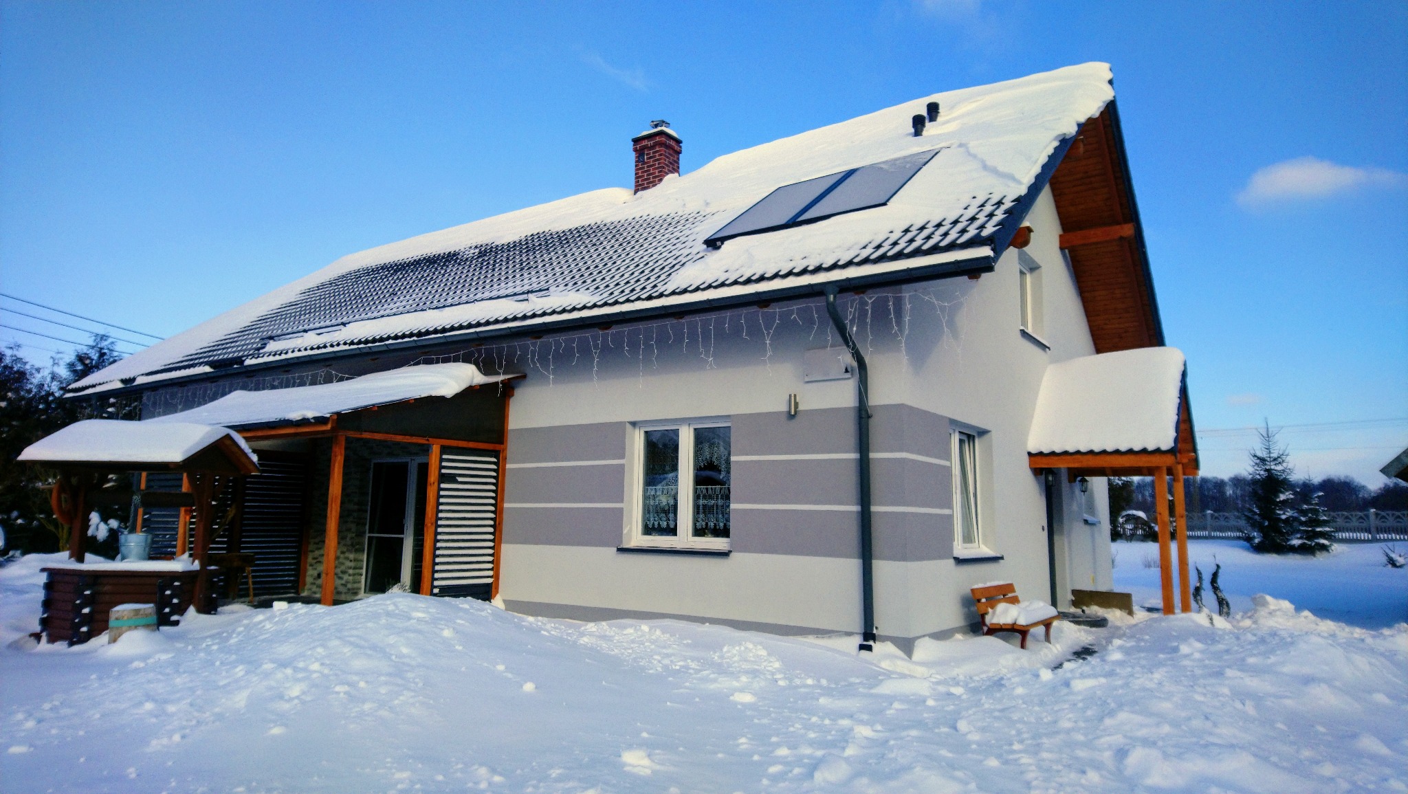 Ośnieżony dom jednorodzinny z szarą elewacją w poziome pasy, panele słoneczne na dachu, studnia z wiadrem i dekoracyjne lampki zwisające z okapu.