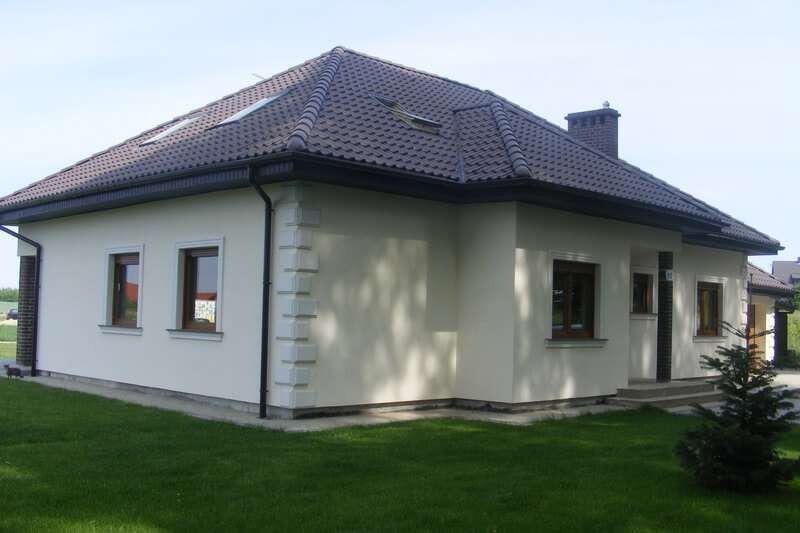 Jednopiętrowy dom z jasną elewacją, ciemnobrązowym dachem z dachówką i ozdobnymi narożnikami elewacji, widziany z poziomu trawnika w słoneczny dzień.