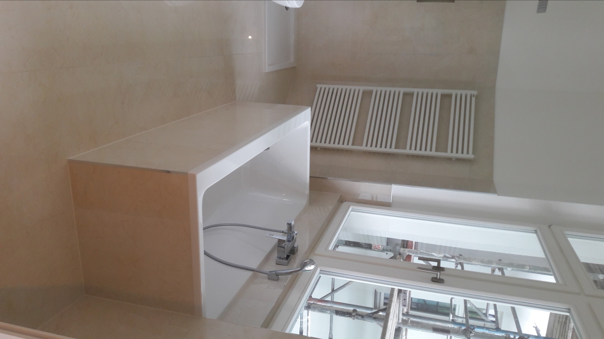 Nowoczesna łazienka z beżowymi płytkami, zabudowaną wanną z chromowaną baterią i grzejnikiem drabinkowym na ścianie, widok z góry na okno z rusztowaniami na zewnątrz.