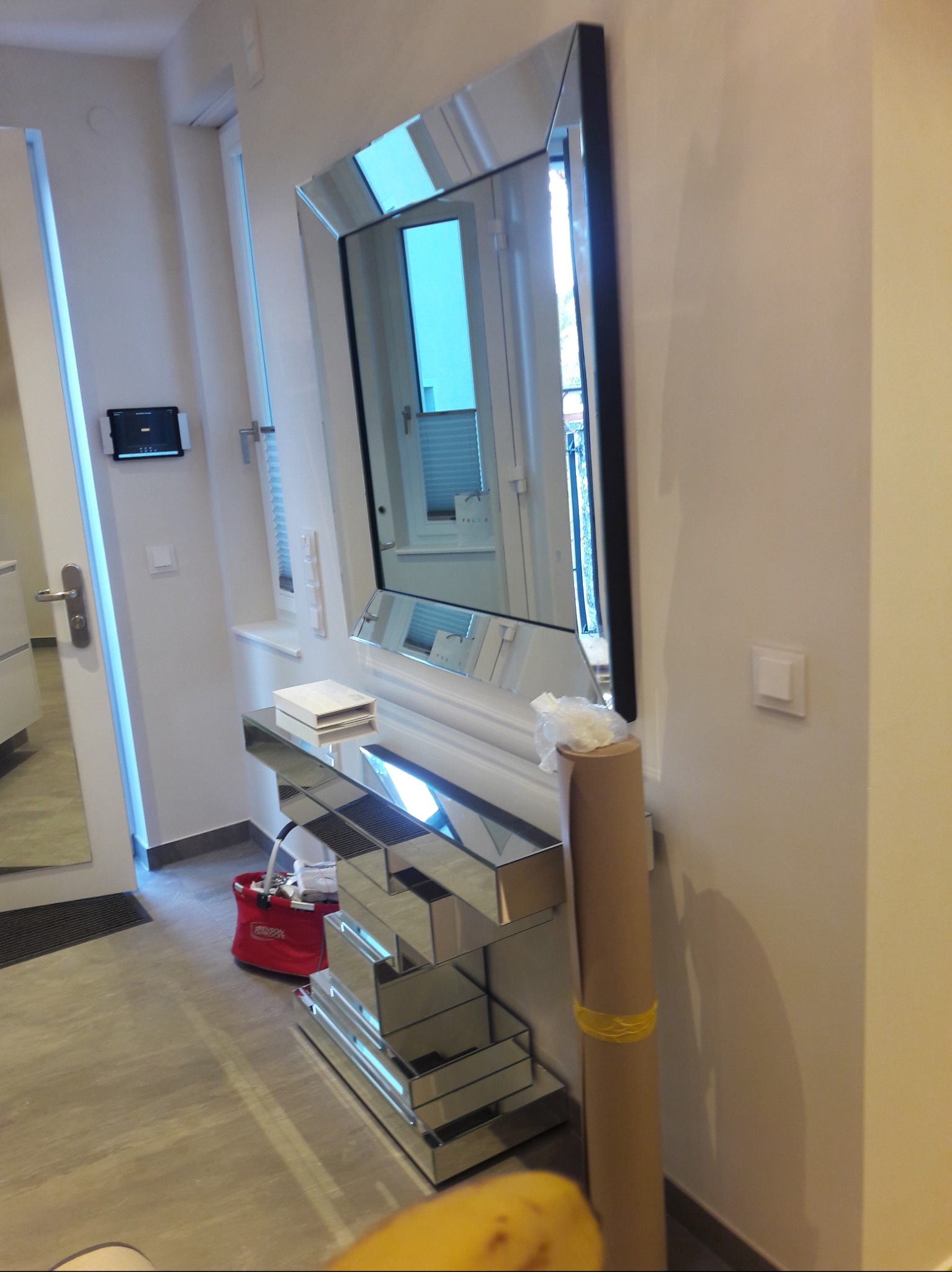 Nowoczesny przedpokój z lustrzaną konsolą i dużym lustrem w ramie, widoczna część wejścia i ściana z zamontowanym tabletem.