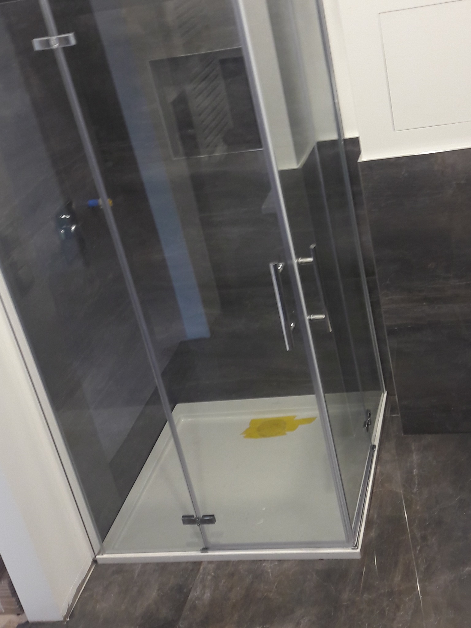 Kabina prysznicowa z przezroczystego szkła z chromowanymi uchwytami, zamontowana na białym brodziku, na tle ciemnych płytek imitujących marmur.