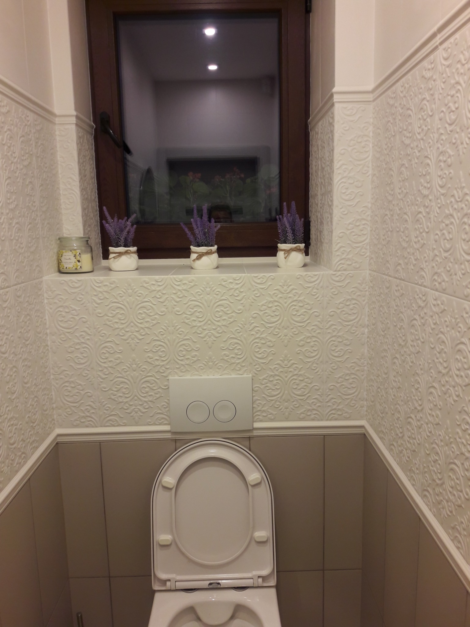 Biała toaleta w małej łazience z dekoracyjnymi, tłoczonymi płytkami ściennymi, okno z kwiatami w tle, trzy doniczki z lawendą i świeczka zapachowa na półce.