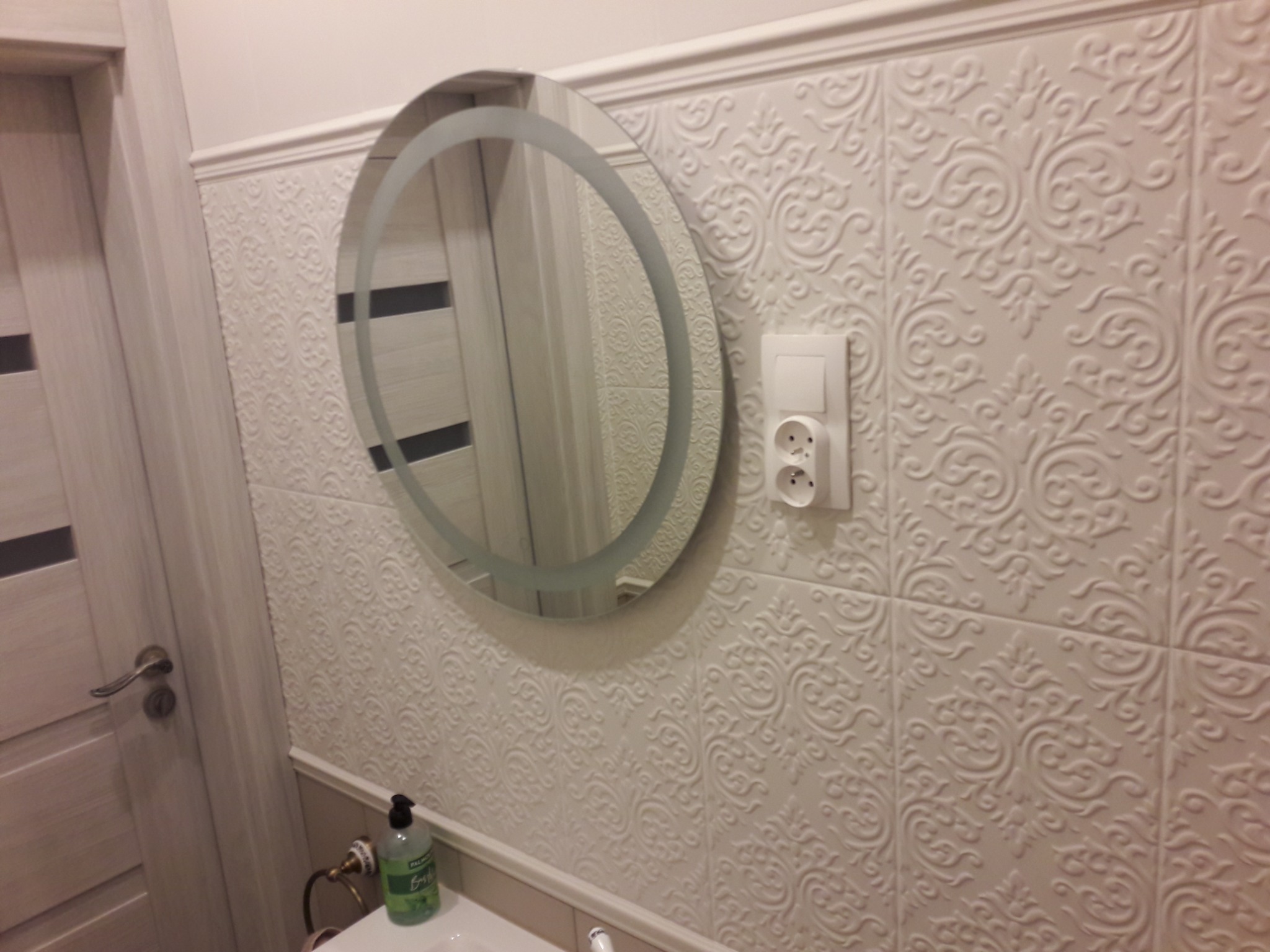 Okrągłe lustro z podświetleniem zawieszone na ścianie z dekoracyjnymi, białymi płytkami. Widoczny fragment umywalki z dozownikiem mydła w płynie oraz drzwi z szarą ramą.