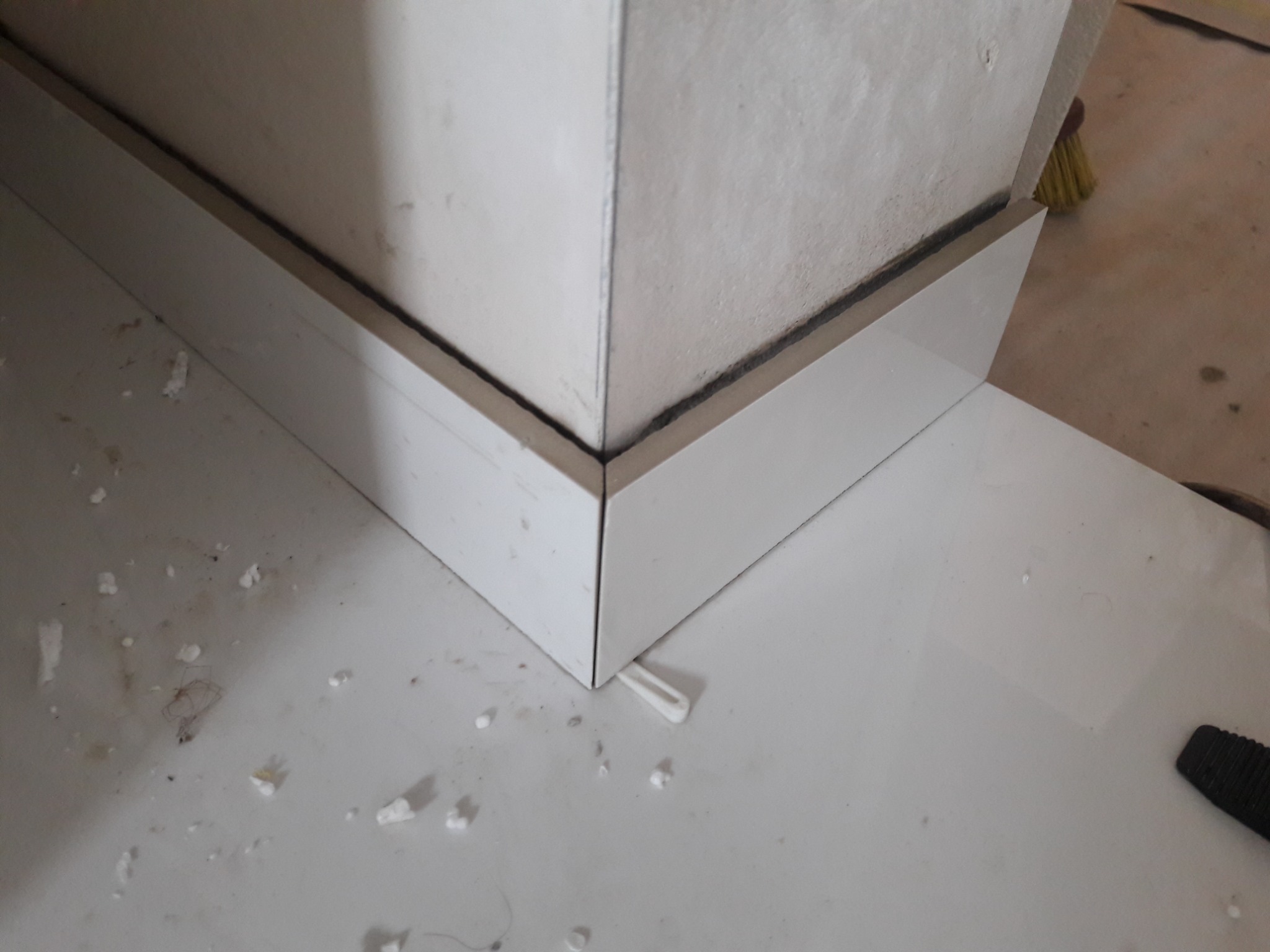 Fragment ściany z zamontowaną białą listwą przypodłogową, widoczne drobne zabrudzenia i resztki materiałów budowlanych na podłodze, narożnik listwy precyzyjnie docięty.