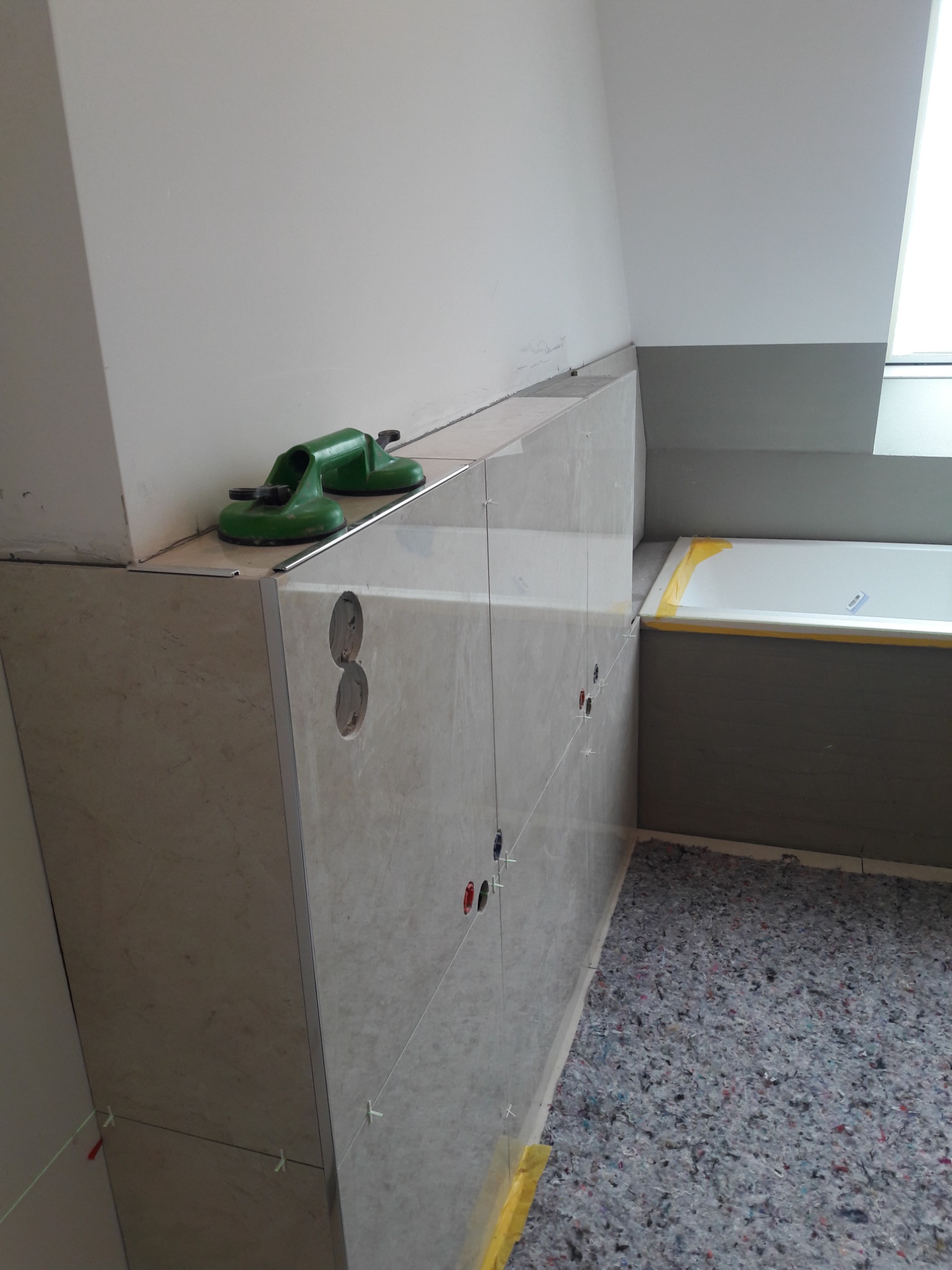 Ściana w łazience w trakcie wykańczania, obłożona beżowymi płytkami imitującymi marmur, z widocznymi otworami na gniazdka elektryczne i krzyżykami dystansowymi; na górze leżą dwie zielone...