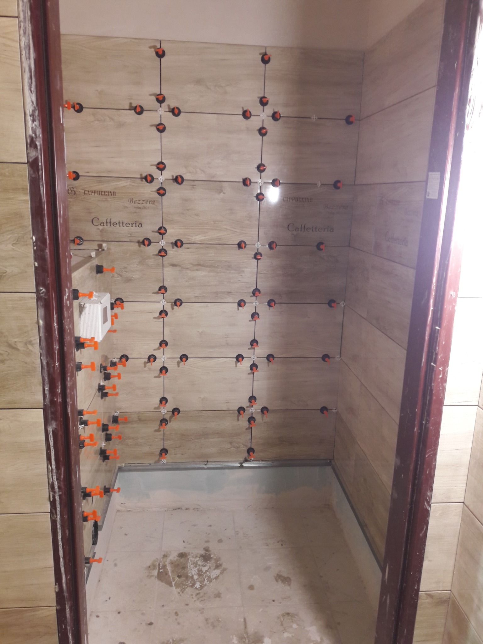 Proces układania płytek imitujących drewno w małej łazience, z użyciem systemu poziomowania płytek z pomarańczowymi klipsami, widoczne napisy na płytkach 'Cappuccino Bezzerra Caffetteria'.