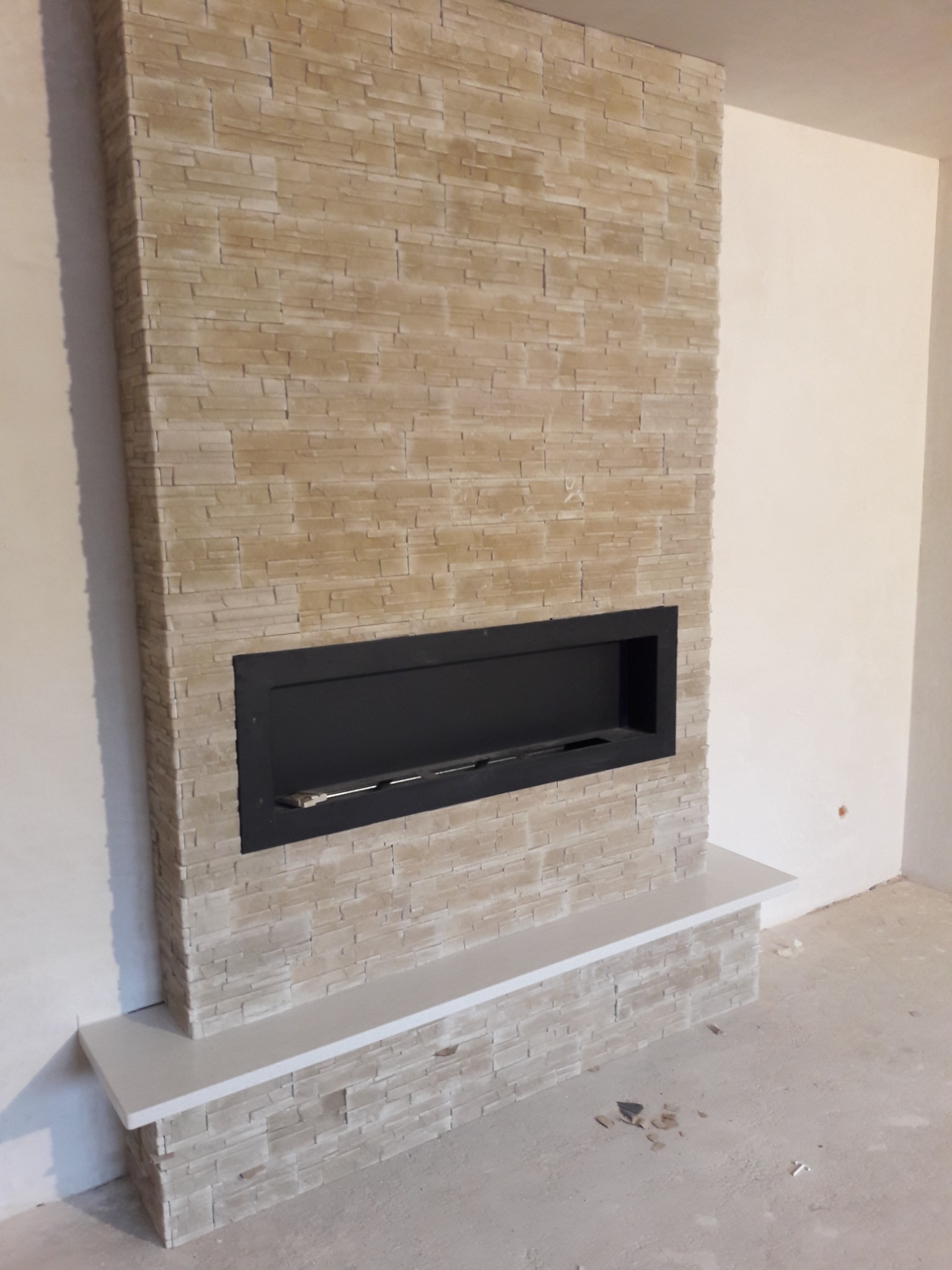 Ściana z okładziną imitującą kamień, z wkomponowanym czarnym biokominkiem i półką, widoczne niedoskonałości podłogi.