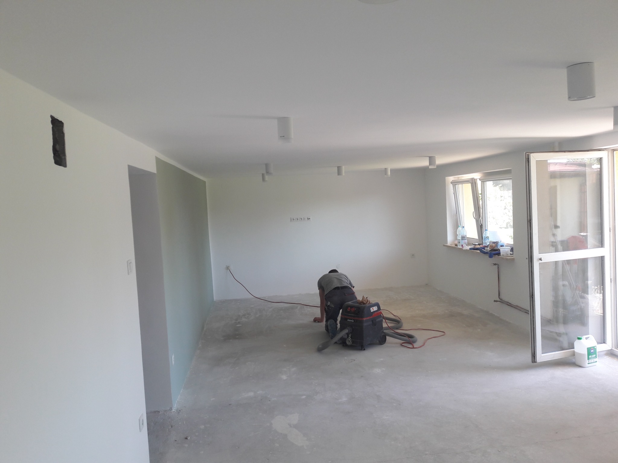 Przygotowanie podłoża pod wylewkę: pracownik szlifuje posadzkę przemysłowym odkurzaczem w jasnym, odnowionym wnętrzu z oknem i białymi ścianami.