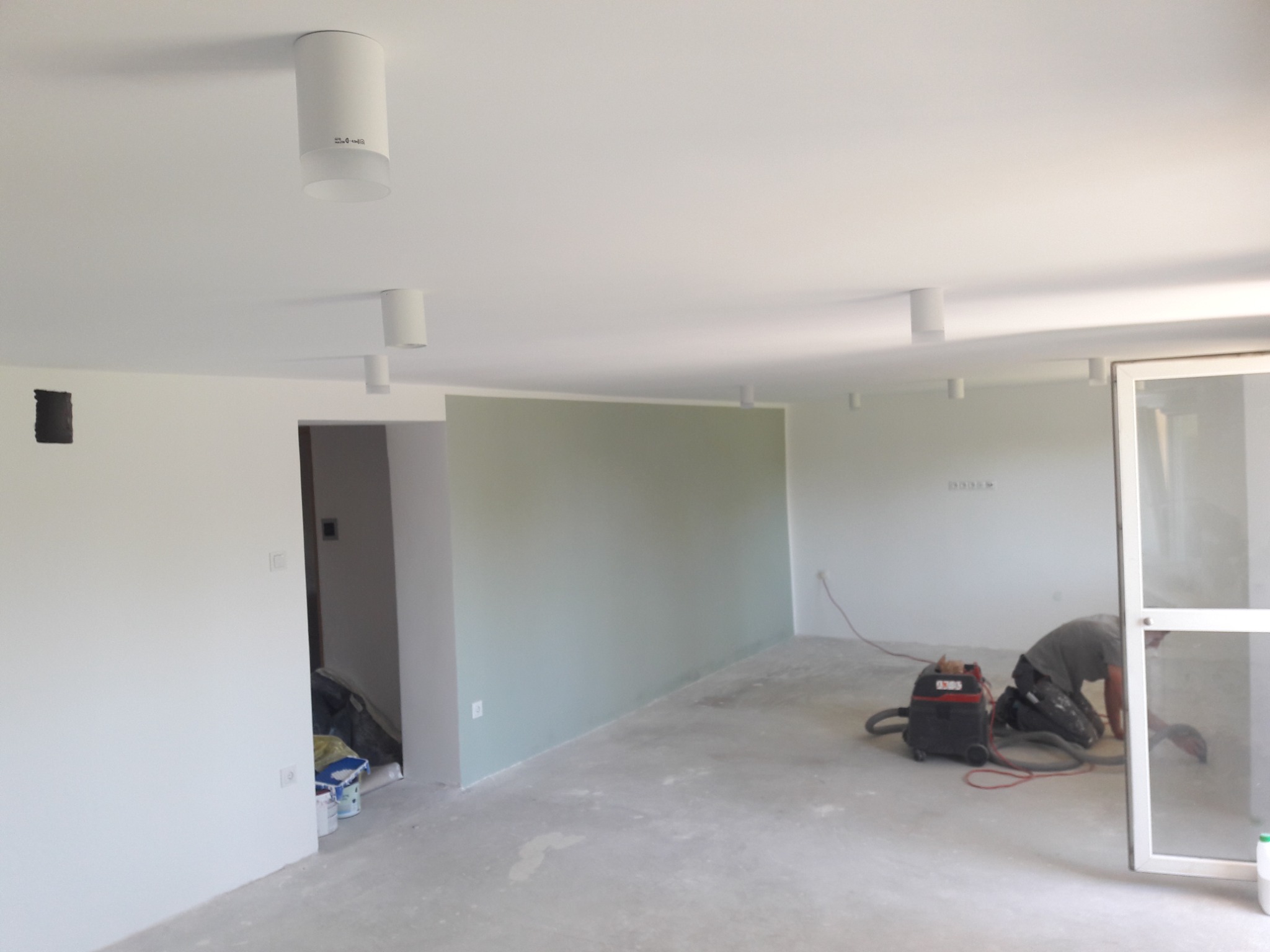 Wnętrze w trakcie prac wykończeniowych: szlifowanie podłogi przy użyciu odkurzacza przemysłowego, widoczne świeżo pomalowane ściany w odcieniach bieli i zieleni, oraz zamontowane cylindryczne lampy...