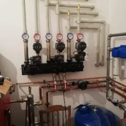 Rozbudowana instalacja centralnego ogrzewania z rozdzielaczem, manometrami i pompami obiegowymi, widoczne rury miedziane i plastikowe, zbiornik wyrównawczy oraz filtry wody, na podłodze leżą...