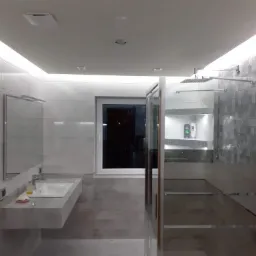 Nowoczesna łazienka w odcieniach szarości z kabiną prysznicową typu walk-in, podświetlanym sufitem i grzejnikiem drabinkowym, widoczna misa umywalkowa z mydłem i dozownikiem.