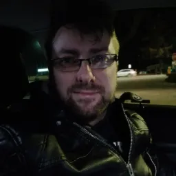 Portret mężczyzny w okularach i czarnej skórzanej kurtce w samochodzie, widok na ulicę z przejeżdżającym białym samochodem w tle.