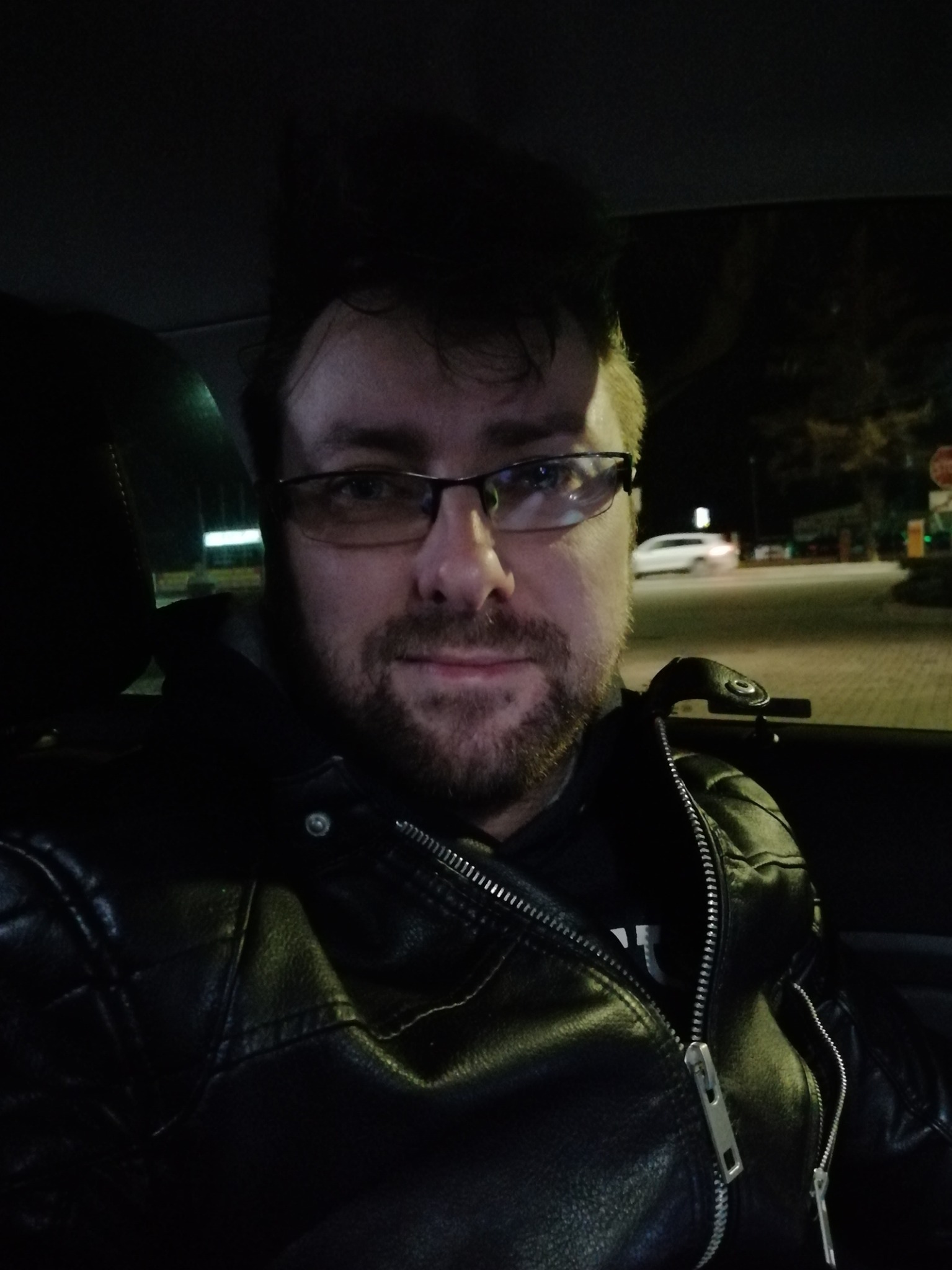 Portret mężczyzny w okularach i czarnej skórzanej kurtce w samochodzie, widok na ulicę z przejeżdżającym białym samochodem w tle.