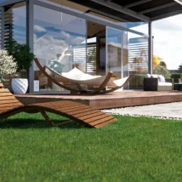 Nowoczesny taras z drewnianym leżakiem na trawie, hamakiem i meblami wypoczynkowymi. Szklana ściana domu odbija błękit nieba. Minimalistyczny design.
