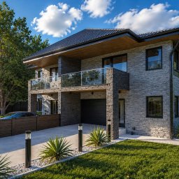 SDesign3D projektowanie wnętrz oraz wizualizacje 3D - Dwukondygnacyjny dom z kamienną elewacją, balkonami z balustradami ze szkła i garażem. Oświetlony podjazd z roślinami i lampami. W tle drzewa i błękitne niebo.