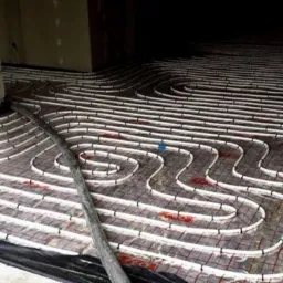 Rozłożona instalacja ogrzewania podłogowego na siatce z folią, widoczne rury w kształcie meandrów, przygotowane do wylewki, z wystającymi kablami i fragmentem ściany z bloczków.