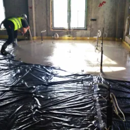 Pracownik w odzieży ochronnej rozprowadza masę wylewkową na podłożu zabezpieczonym folią, widoczne okna i narzędzia pomiarowe.