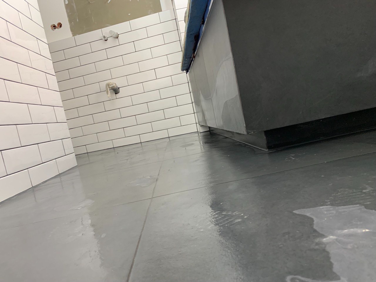 Łazienka w trakcie remontu: białe, prostokątne płytki na ścianach, szare płytki podłogowe, fragment czarnej szafki pod umywalkę, widoczne ślady wody na podłodze.