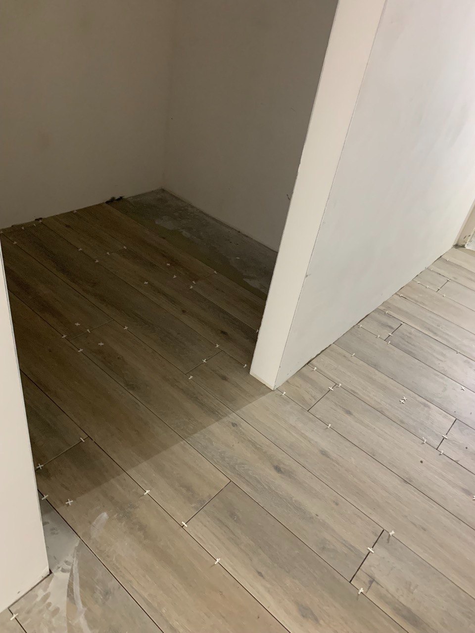 Układanie płytek imitujących drewno w małym pomieszczeniu, widoczne białe krzyżyki dystansowe, ściany przygotowane do dalszych prac wykończeniowych.