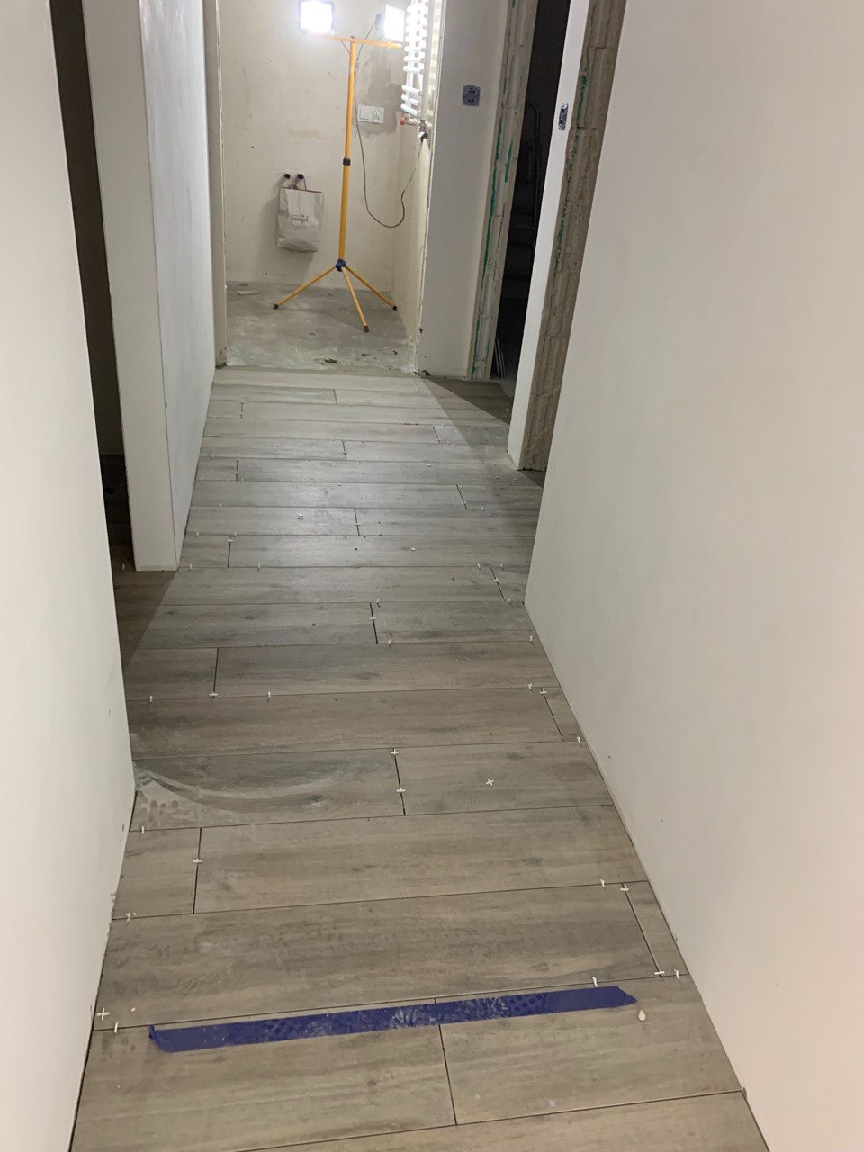 Korytarz w trakcie układania podłogi z płytek imitujących drewno; widoczne krzyżyki dystansowe i taśma malarska, w tle żółty statyw z lampą budowlaną.