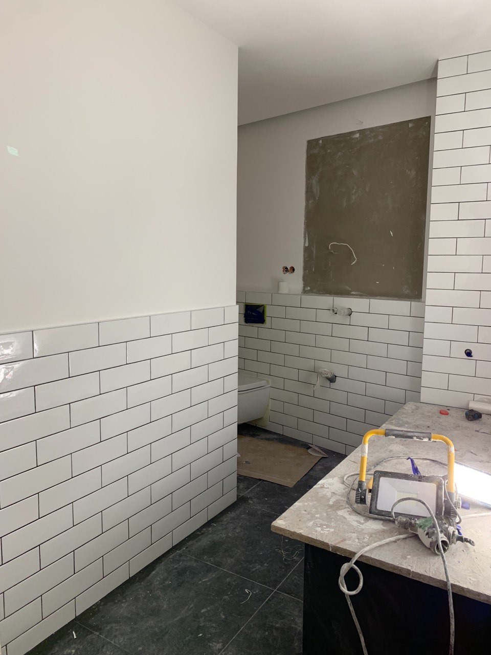 Białe, prostokątne płytki ułożone na ścianie w łazience w trakcie remontu, widoczny fragment toalety i lampa budowlana na blacie, ściana z szarym fragmentem przygotowanym pod lustro.