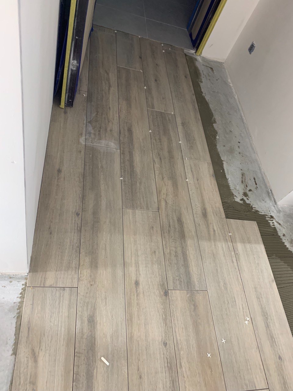 Układanie podłogowych płytek imitujących drewno w trakcie prac, widoczne krzyżyki dystansowe i świeży klej przy ścianie.