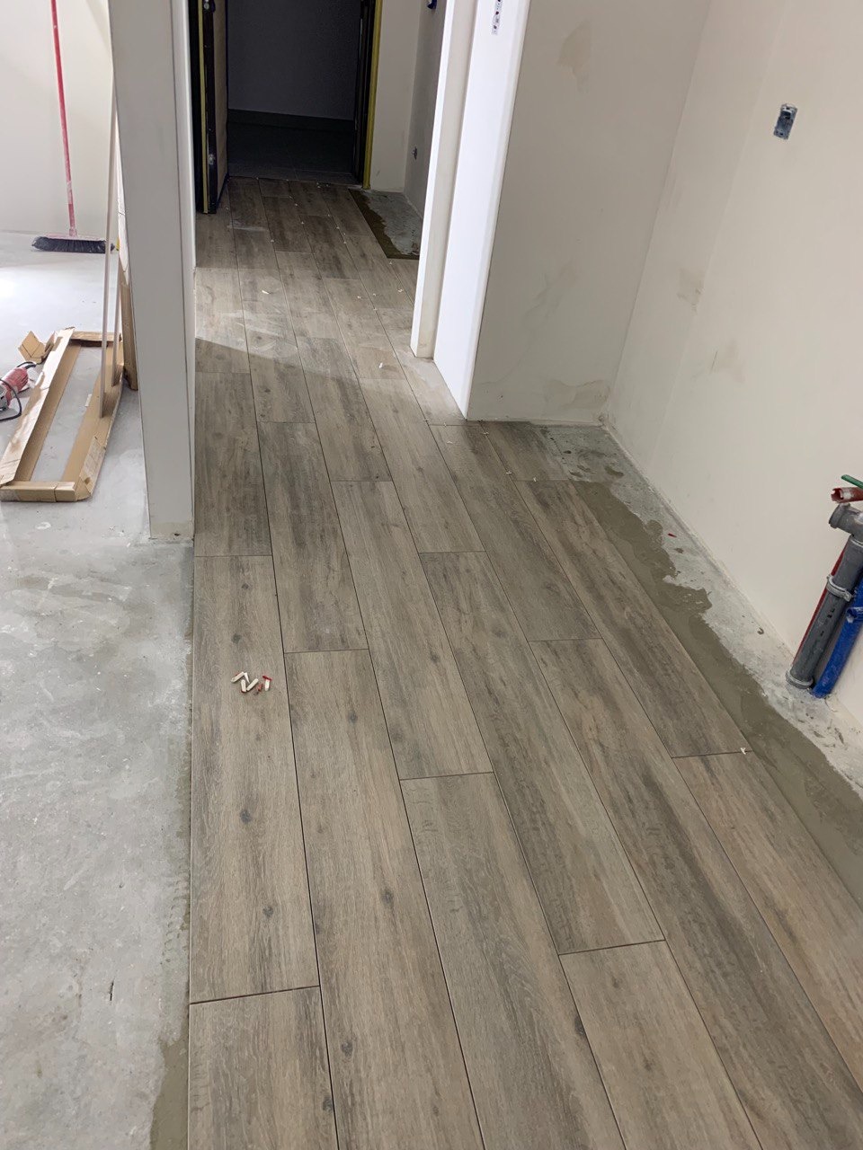 Korytarz w trakcie układania podłogi z płytek imitujących drewno, widoczne niedociągnięcia w postaci niezaspoinowanych przestrzeni przy ścianach i resztki zaprawy na podłodze.