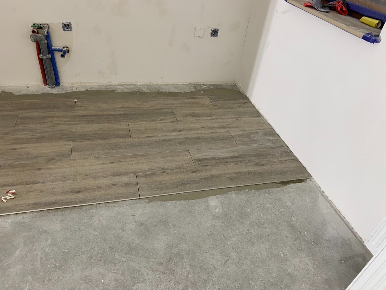 Układanie podłogi z płytek imitujących drewno w trakcie remontu, widoczne rury instalacyjne na ścianie i zapasowe krzyżyki dystansowe.