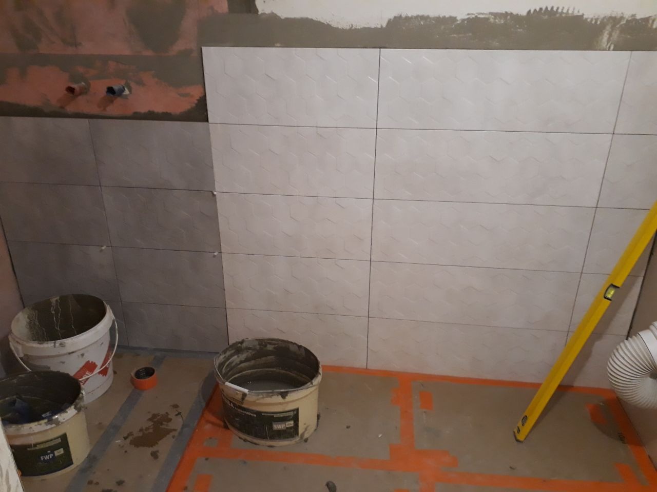Ściana łazienki w trakcie układania płytek: po lewej stronie szare płytki, po prawej beżowe z delikatnym wzorem geometrycznym, widoczne dwa wiadra z zaprawą, poziomica przy ścianie, pomarańczowe...
