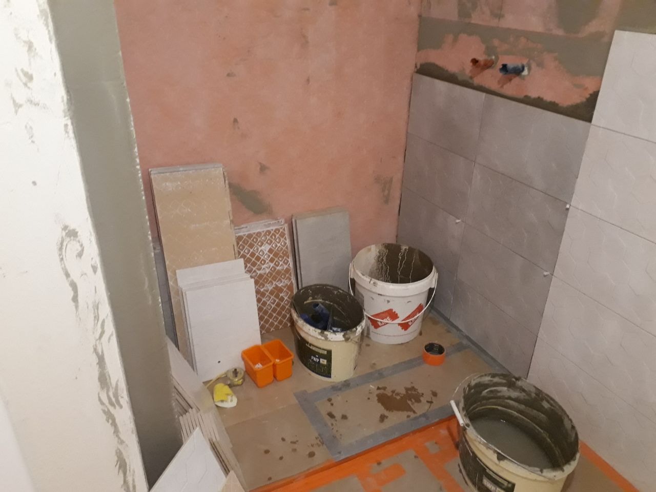 Prace glazurnicze w trakcie realizacji w łazience w Warszawie: widoczne ułożone płytki na ścianie, zaprawa w wiadrach i zapas płytek podłogowych.