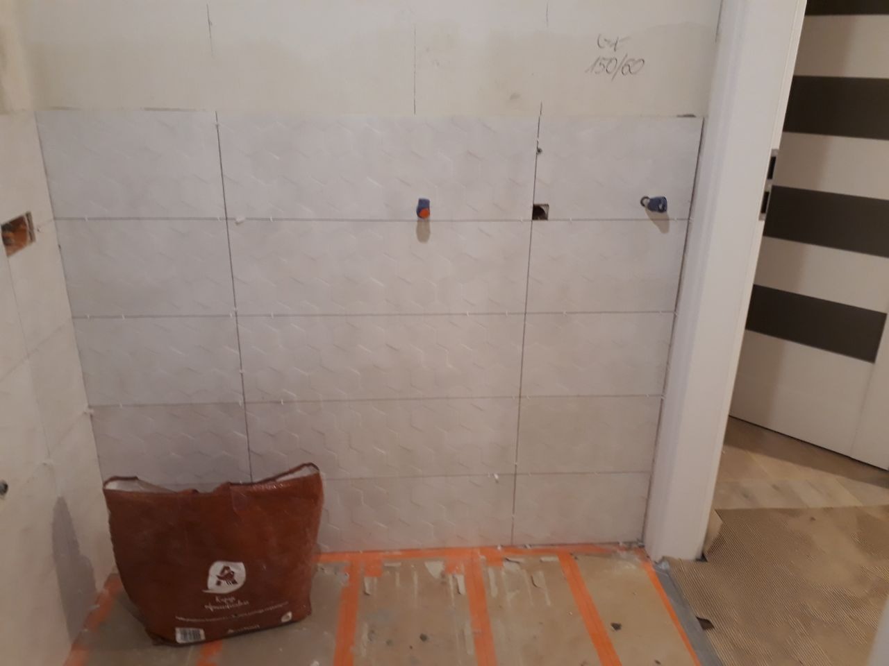 Ściana łazienki w trakcie układania jasnych, wzorzystych płytek ceramicznych o wymiarach 150/60, widoczne krzyżyki dystansowe, przygotowane otwory na instalacje i worek z zaprawą klejową na podłodze.