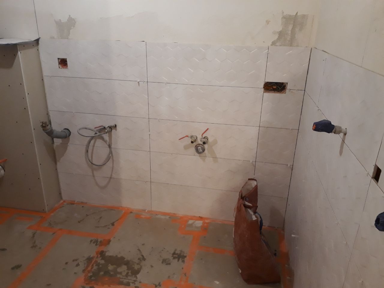 Białe, wzorzyste płytki ułożone na ścianie łazienki w trakcie remontu, widoczne wystające rury i instalacje, pomarańczowe linie wyznaczające obszar układania płytek na podłodze, worek z zaprawą...