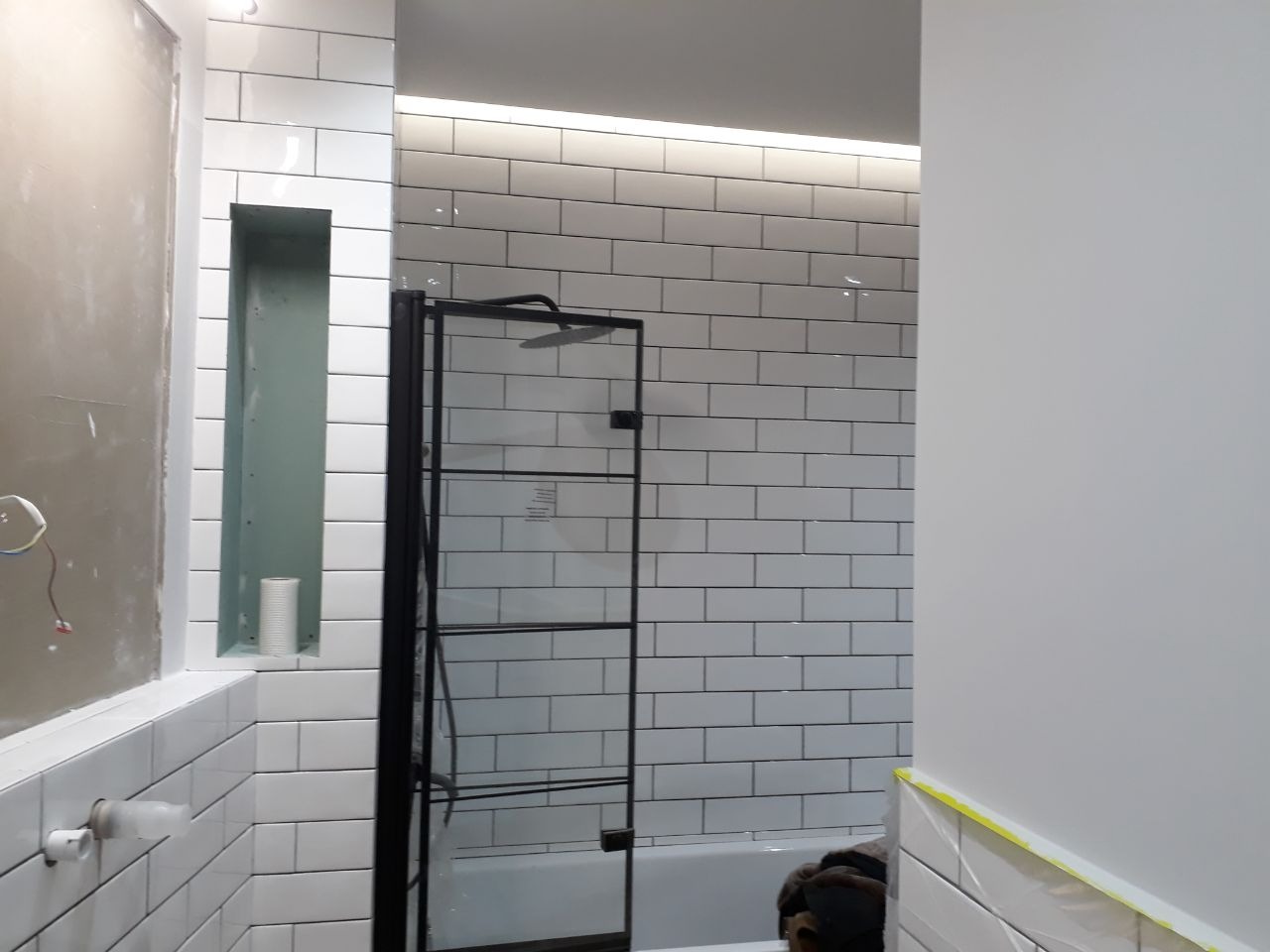 Niedokończona łazienka z białymi, prostokątnymi płytkami ułożonymi jak cegły, czarną kabiną prysznicową i wnęką w ścianie, w której stoi rolka papieru toaletowego.