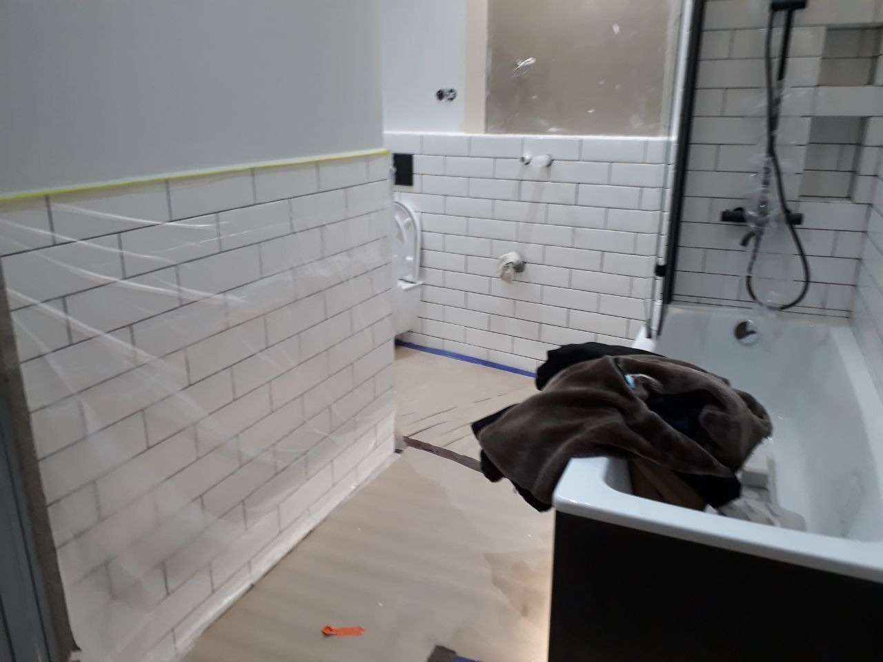 Łazienka w trakcie remontu: białe, prostokątne płytki na ścianach, zabezpieczone folią i taśmą malarską, wanna z czarną obudową, częściowo zasłonięta stertą ubrań. Widoczne niedokończone prace przy...