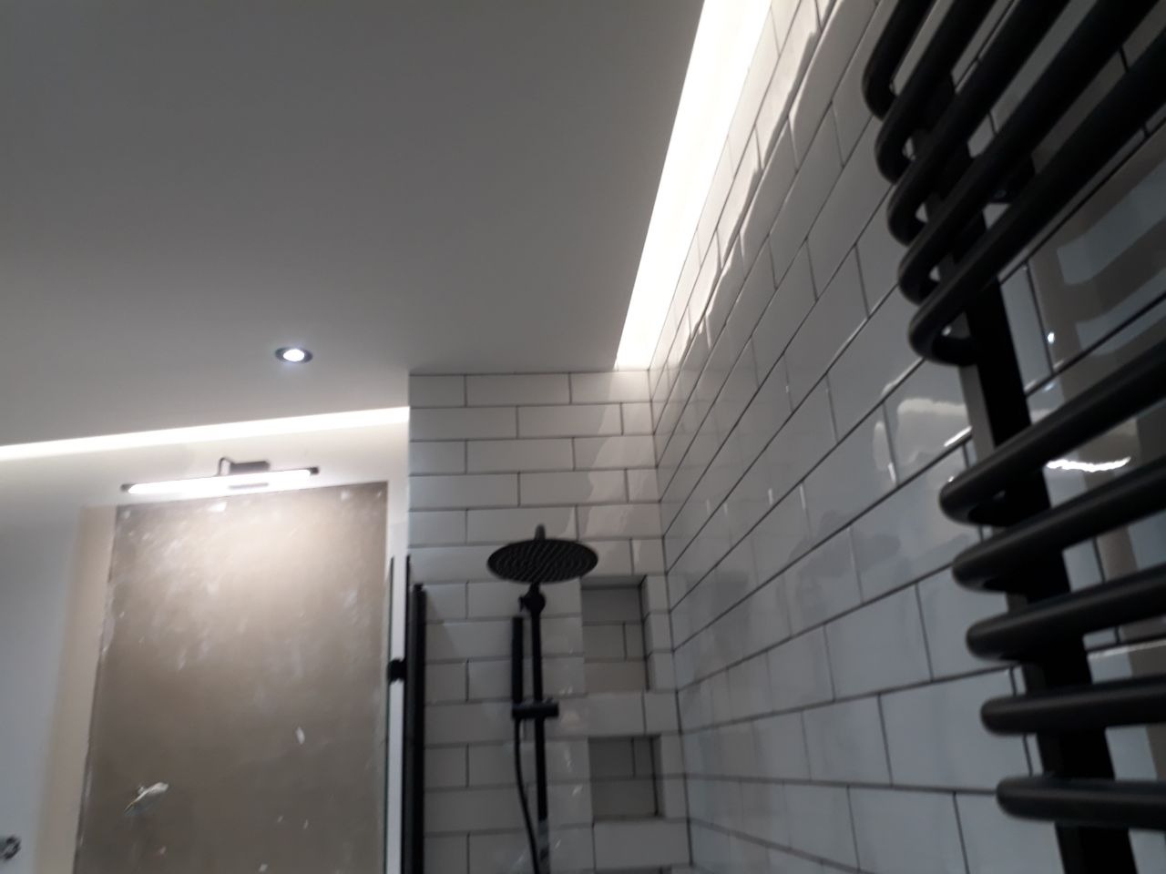 Nowoczesna łazienka w trakcie wykańczania: białe prostokątne płytki na ścianie, czarna deszczownica i grzejnik drabinkowy, sufit podwieszany z oświetleniem LED, ściana przygotowana do malowania.